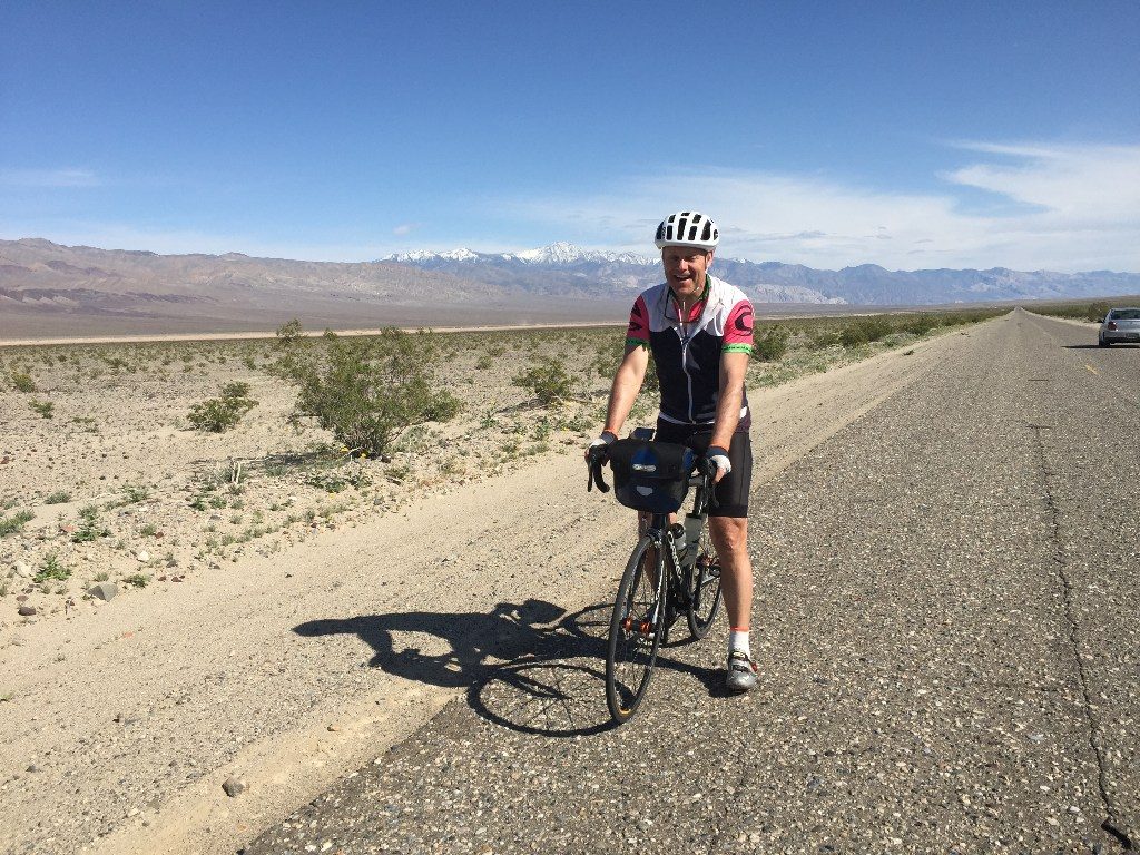 Chris cycling in Death Valley California, on a long strip of road on the valley floor, snow capped mountains in the background