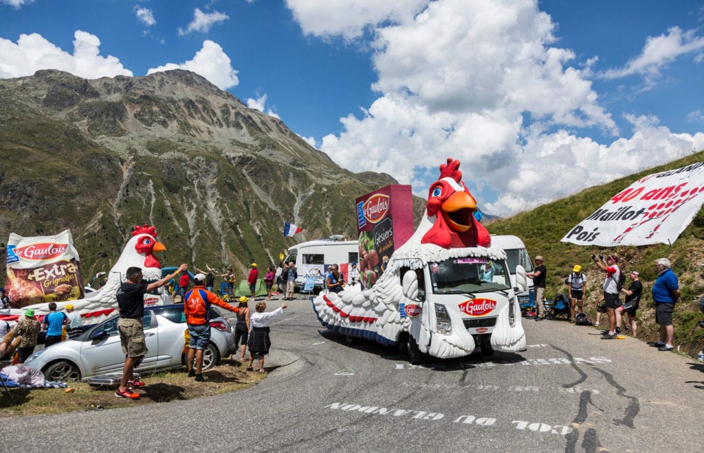 photo of a motorized float in the tour de france caravan, featuring oversize roosters to promote chicken nuggets, winding up a mountain road, 