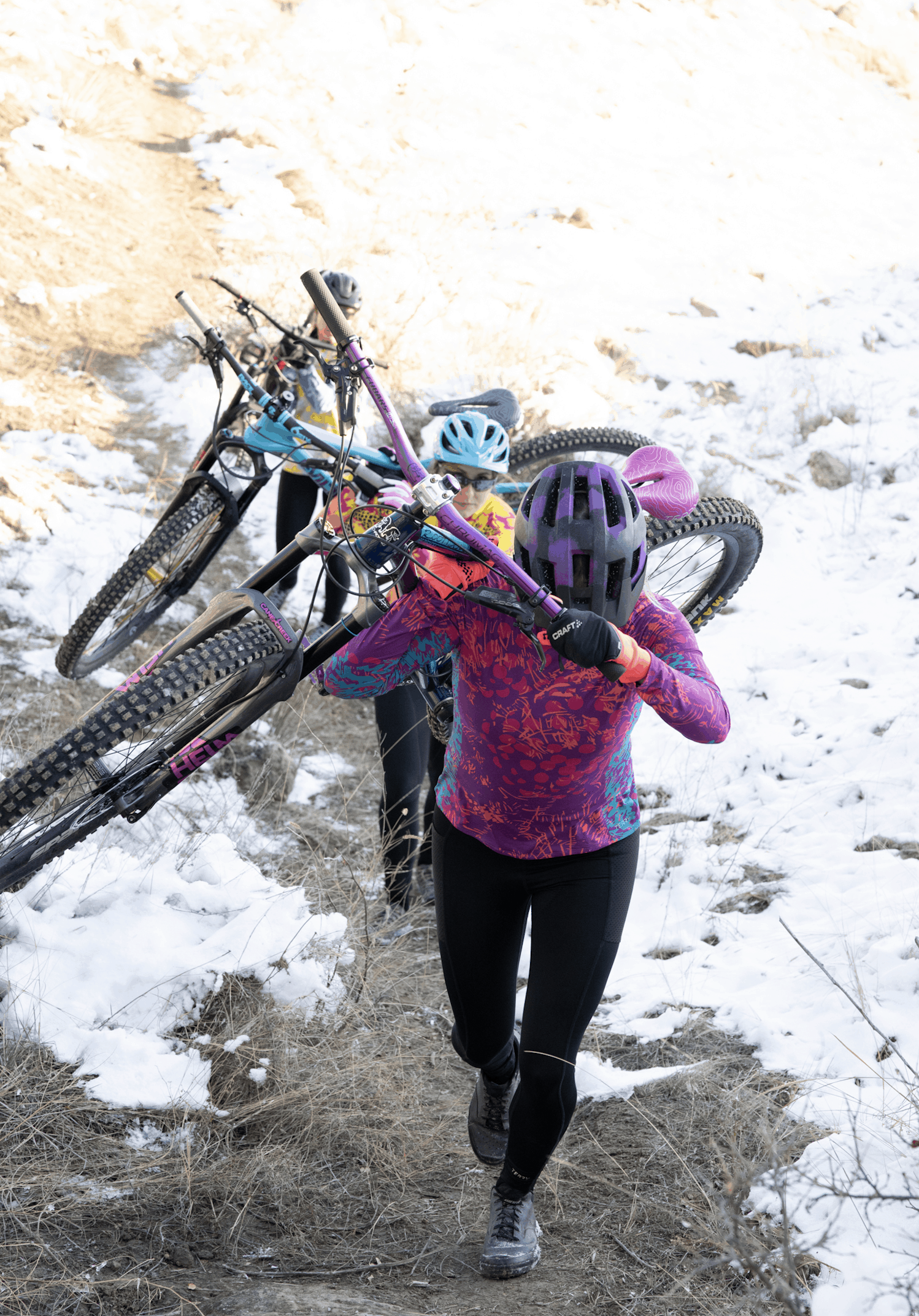Women shouldering their mountain bikes to climb a hill, snow on ground, wearing Terry bike jerseys and cycling tights