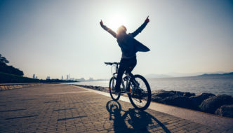 Woman cycling alongside a river, cityscape in the far distance, holding arms up joyfully