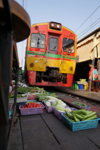 A train makes its way along a narrow street lined with market stalls and produce, pulled quickly out of the way as the train passes.
