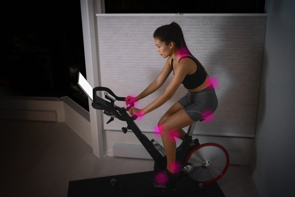 Woman riding an indoor exercise bike or peloton bike, with highlights showing pain points from incorrect riding position
