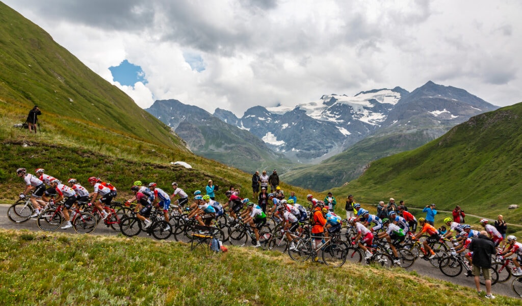 Peloton rides up a mountain road with soaring peaks in teh background, Tour de France 2019